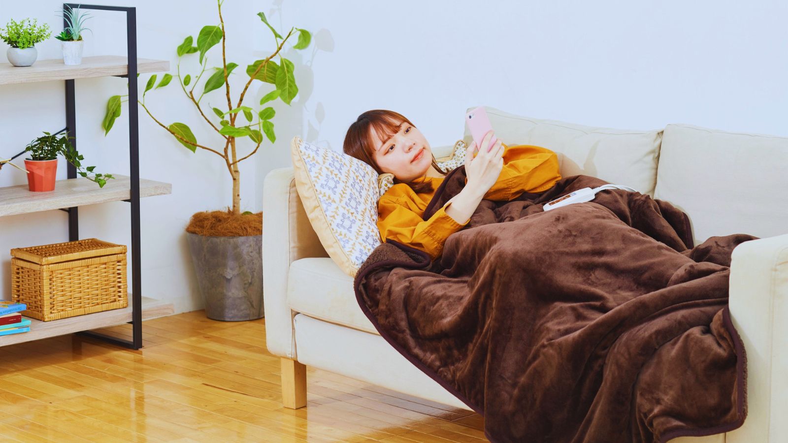 A person sitting on a couch with a blanket covering them, holding a phone, with a calm but pensive expression.