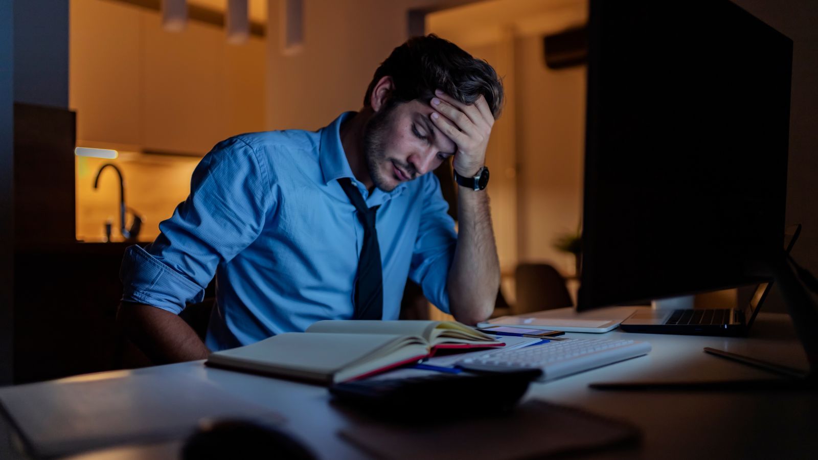 A person sitting at a cluttered desk with their head resting on one hand, conveying the weight of ongoing low-grade stress.