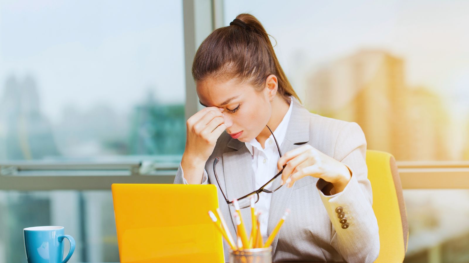 A woman at an office desk pinching the bridge of her nose, glasses in her other hand, in front of an open laptop.