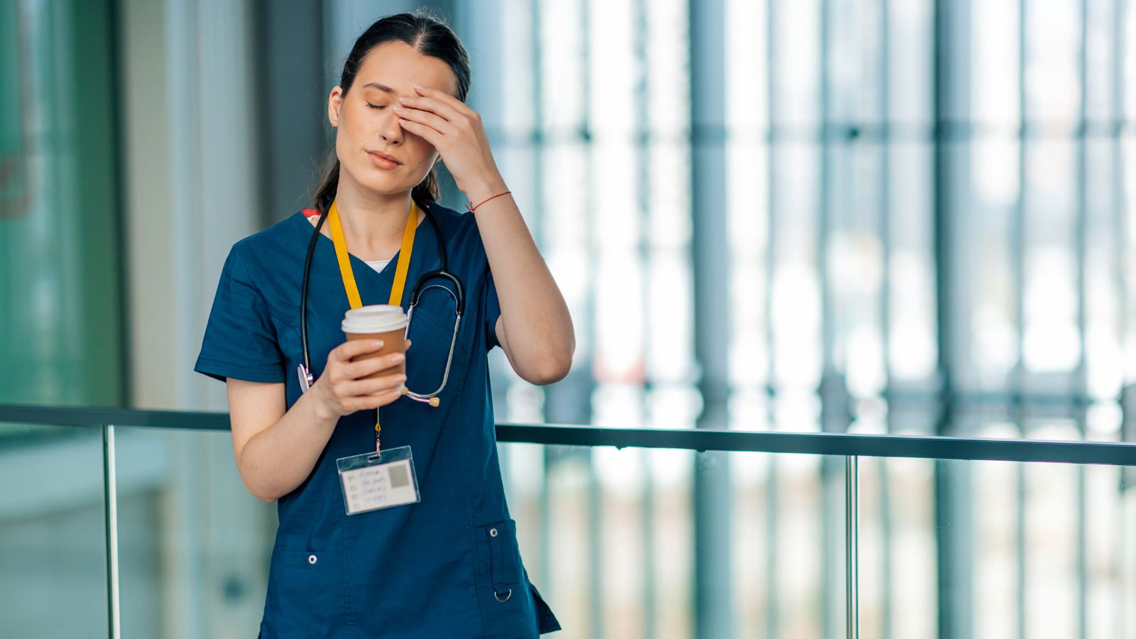 A nurse wearing scrubs, holding a coffee, with her eyes closed and a hand on her head,looking burnt out.