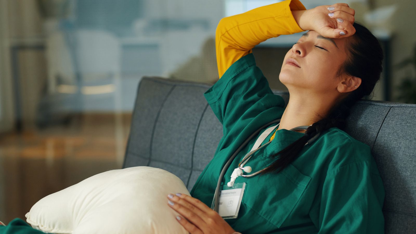 A nurse in scrubs sitting on a couch looking tired and stressed