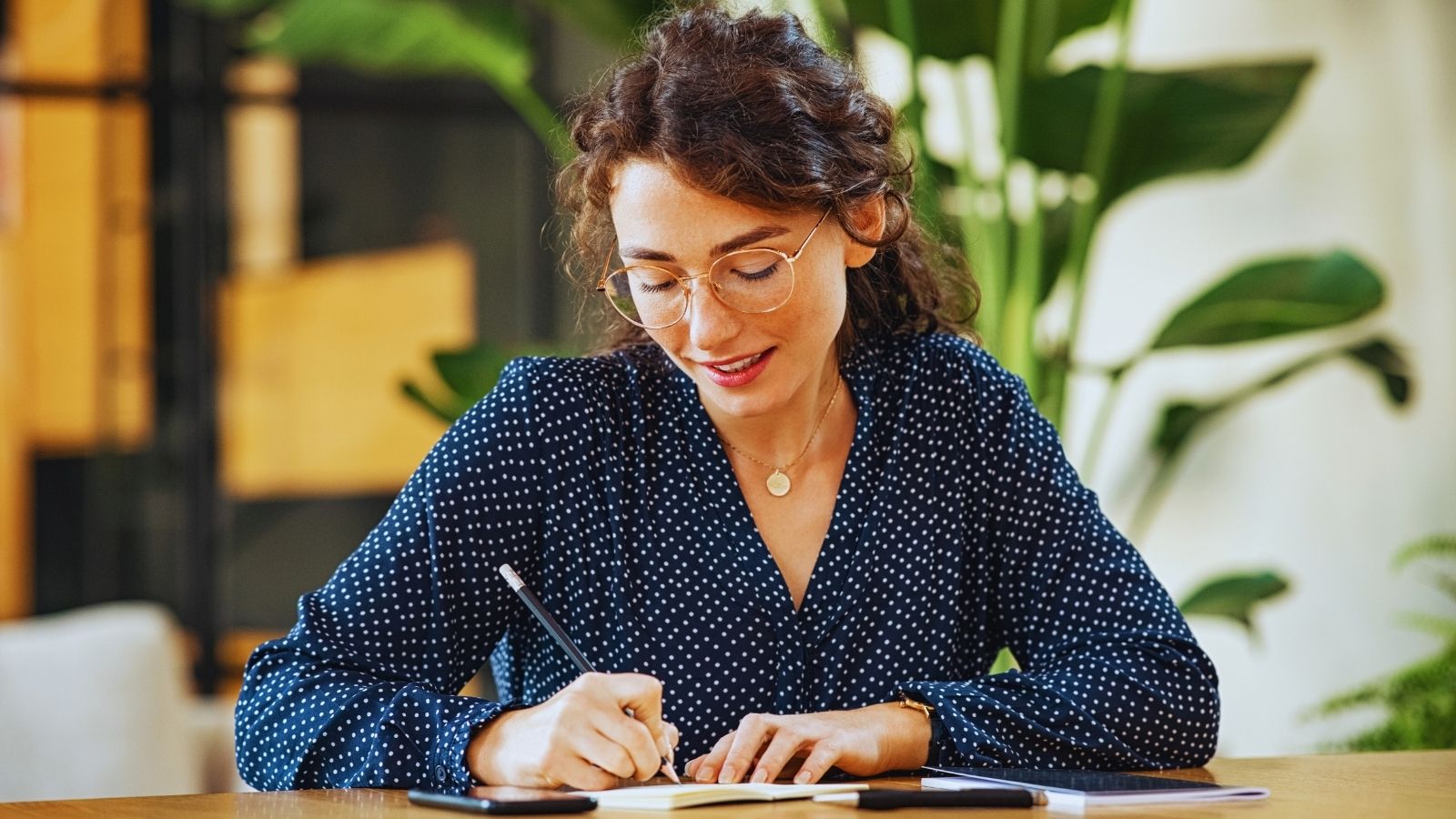 A woman writing a short entry in a notebook at a table, with her phone face down beside her.