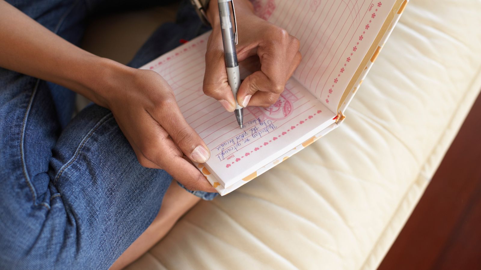A close-up of a hand writing short structured entries in a small notebook on a wooden desk.